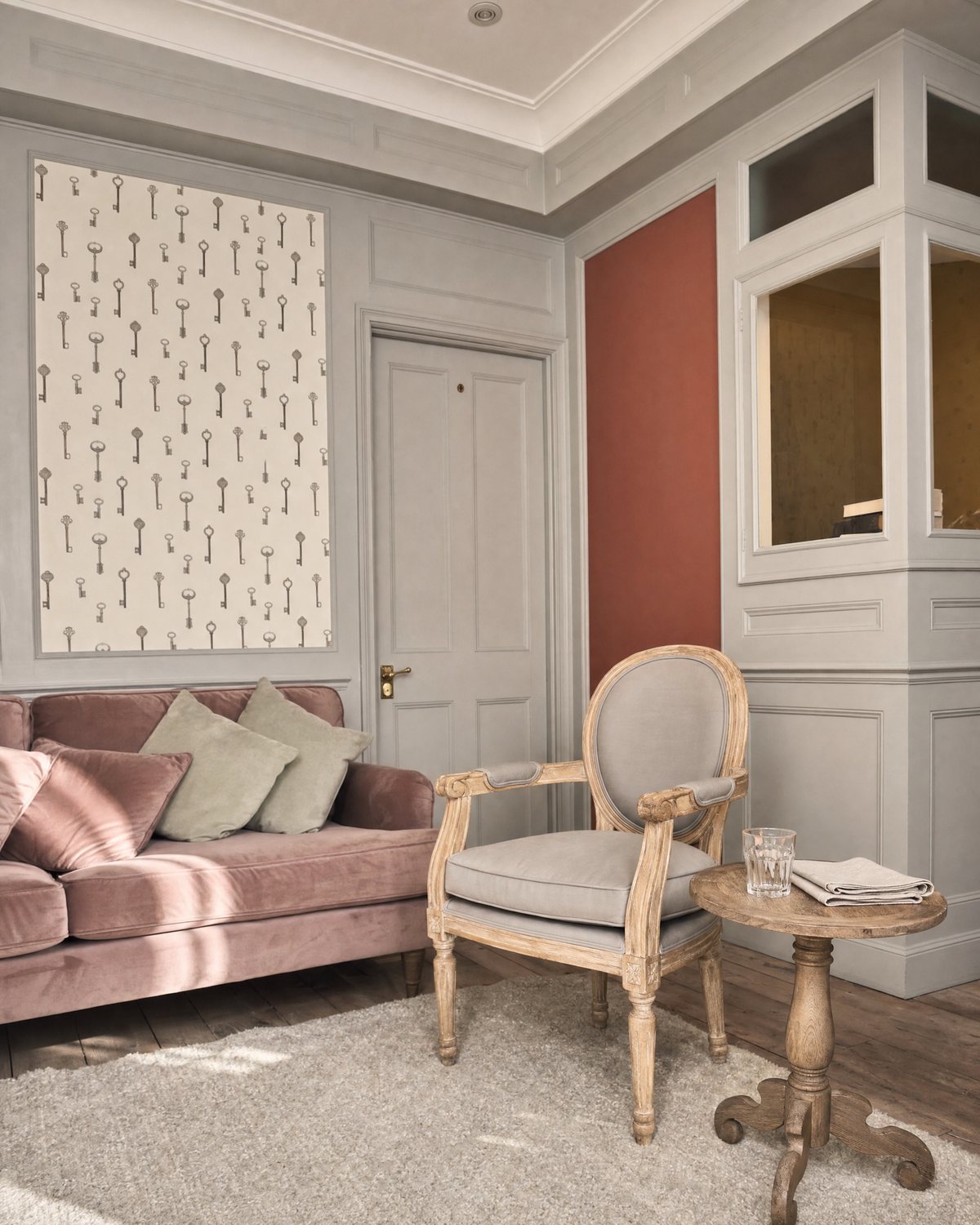 A quiet consulting room with a pink velvet sofa, an oak armchair and key-pattern wallpaper